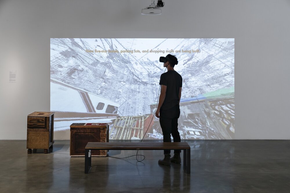 Building the City of Exiles, San Francisco Arts Commission, 2018. Sculpture, video, and virtual reality. A person wearing a virtual reality headset stands in front of a projection of a skyline of scaffolds with an unhoused encampment in the foreground. A caption above the image reads "New five-star hotels, parking lots, and shopping malls are being built."