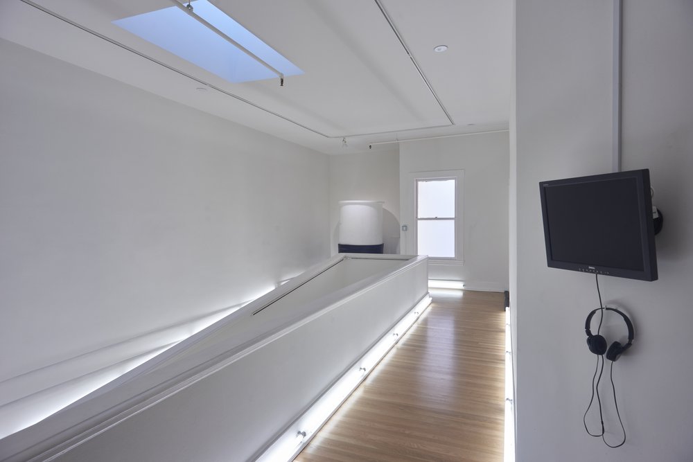 ALBEDO, 2021 An art gallery with white walls, a wood floor bordered by fluorescent lights, and a small display screen with headphones.