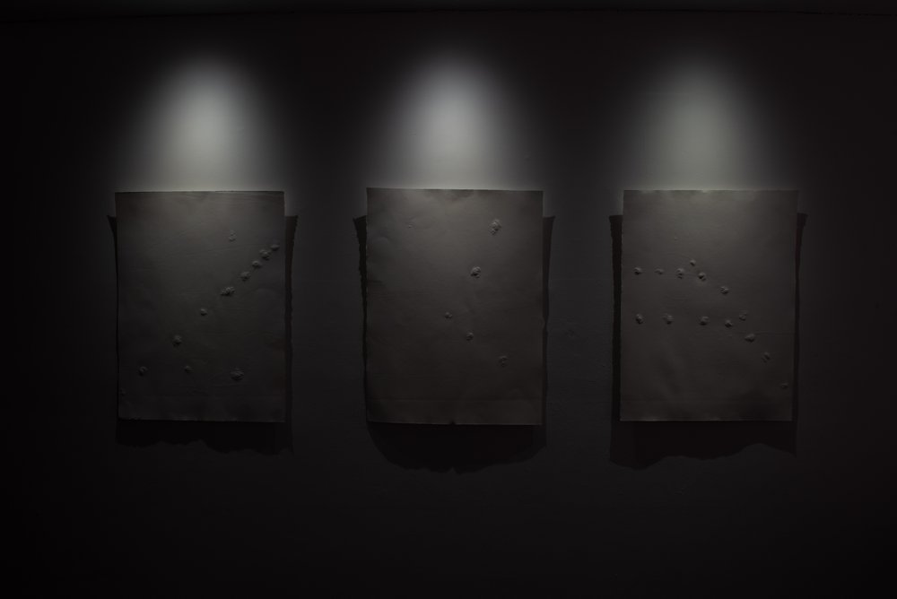 Ghatel, 2021 A dark gallery wall with three illuminated canvases displaying different arrangements of embossed marks.