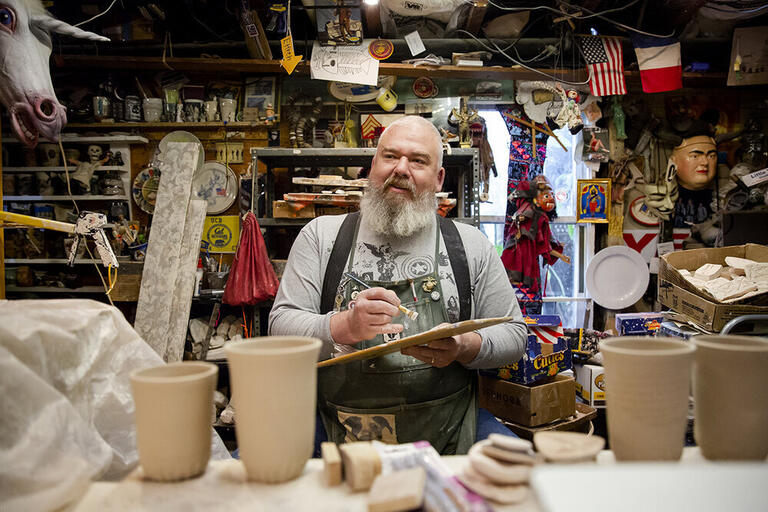 A portrait of Ehren Tool in a ceramics studio. He is holding a paintbrush and palette, and he is surrounded by various art projects.